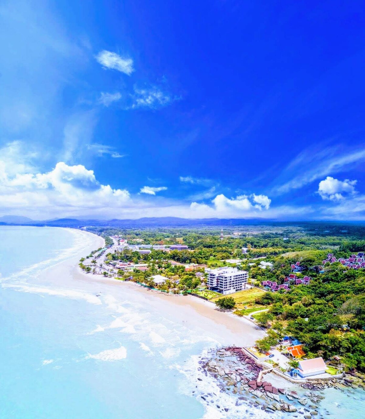 An expansive view captures the coastline with sandy beaches meeting the turquoise sea. Lush greenery and residential areas line the shore, with gentle waves reaching the beach. A clear blue sky, dotted with white clouds, stretches overhead, enhancing the serene atmosphere of the locale.