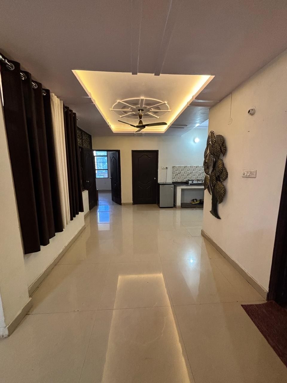 A hallway with glossy tiled flooring is illuminated by ceiling lights. The walls are adorned with dark curtains, and a modern ceiling fan is positioned above. An open doorway leads to a kitchenette, while a decorative wall feature adds interest to the space.