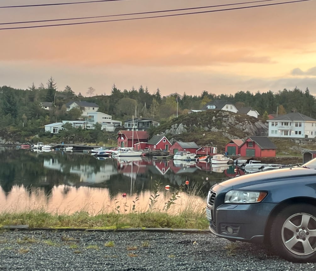 A serene waterfront scene is captured at sunset, where colorful houses are reflected in calm waters. Various boats are anchored nearby, and a parked car is positioned in the foreground. Lush greenery and a rocky hillside add to the tranquil landscape.