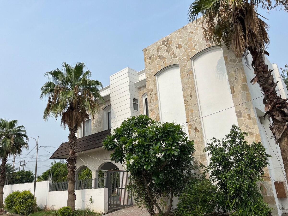 The villa is showcased with a modern stone facade, complemented by lush palm trees and vibrant greenery. A spacious entrance is visible, providing access to the serene garden area, while the sky above reflects a clear blue.
