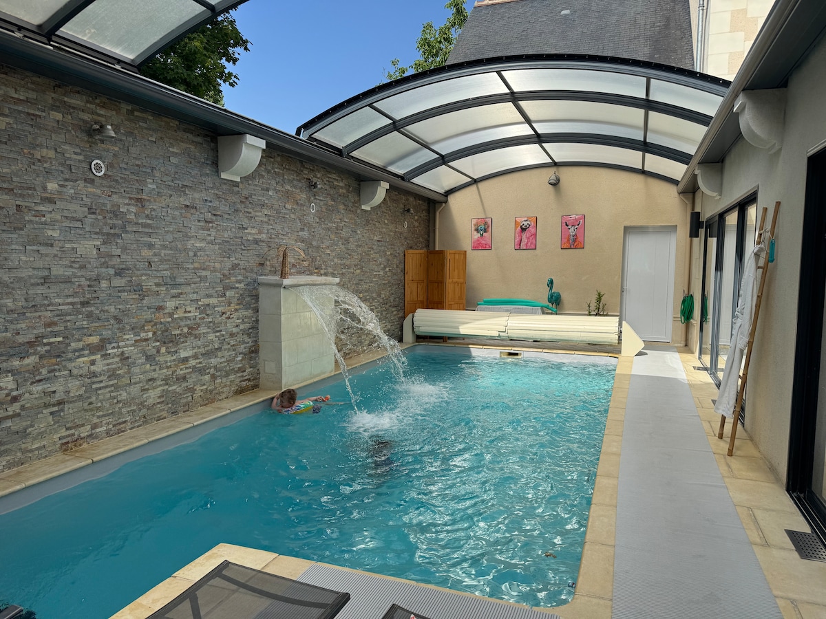 An indoor pool area is featured, with a clear retractable roof allowing natural light to flood in. The pool includes a gentle fountain and is surrounded by stone walls. A lounge area with a covered seating section is visible, providing a tranquil setting.