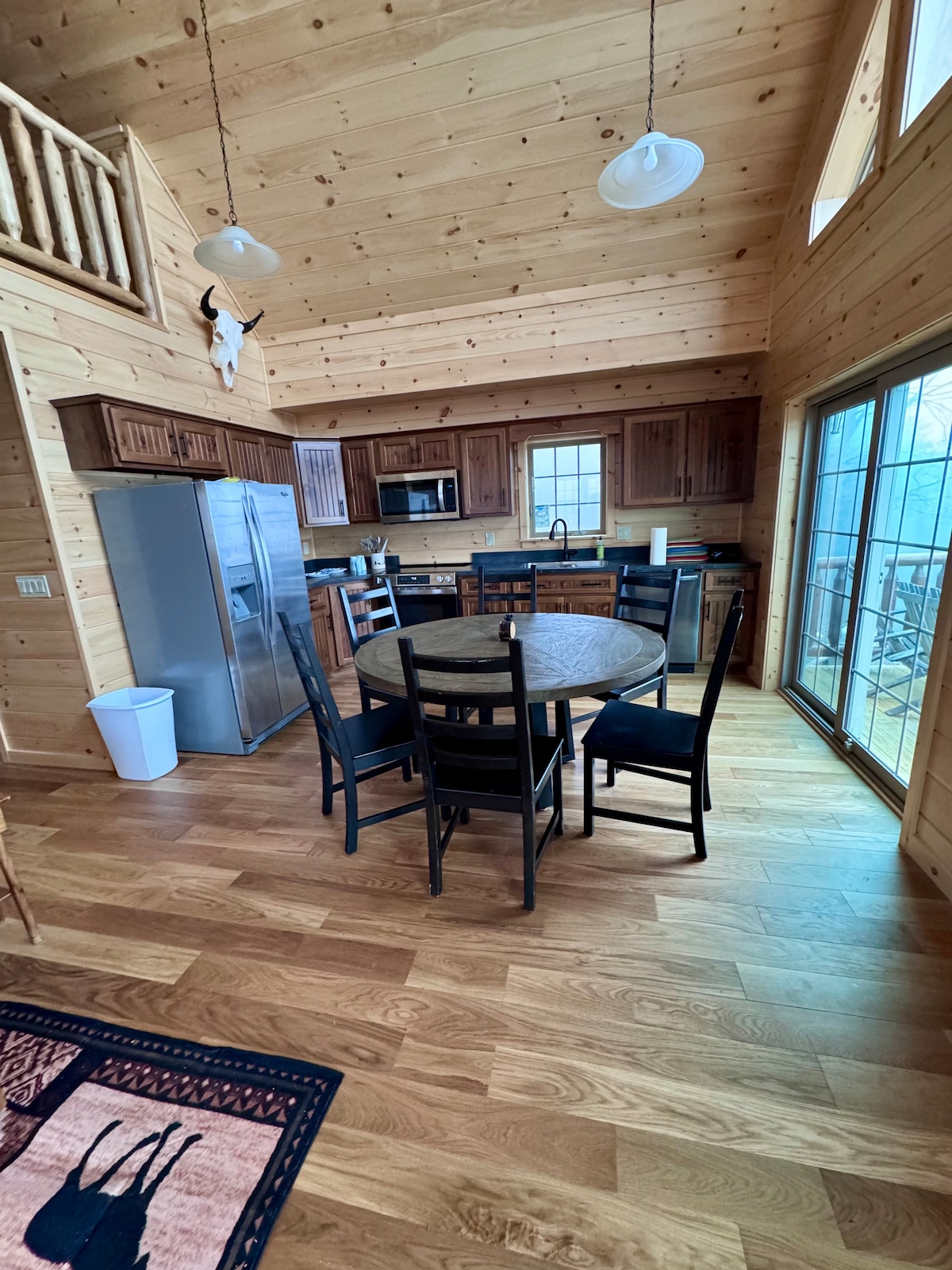The kitchen area features wooden cabinetry and modern appliances, including a refrigerator and microwave. A round dining table with black chairs is positioned near large windows, providing ample natural light. The wood flooring adds warmth to the spacious room, enhancing the overall aesthetic.
