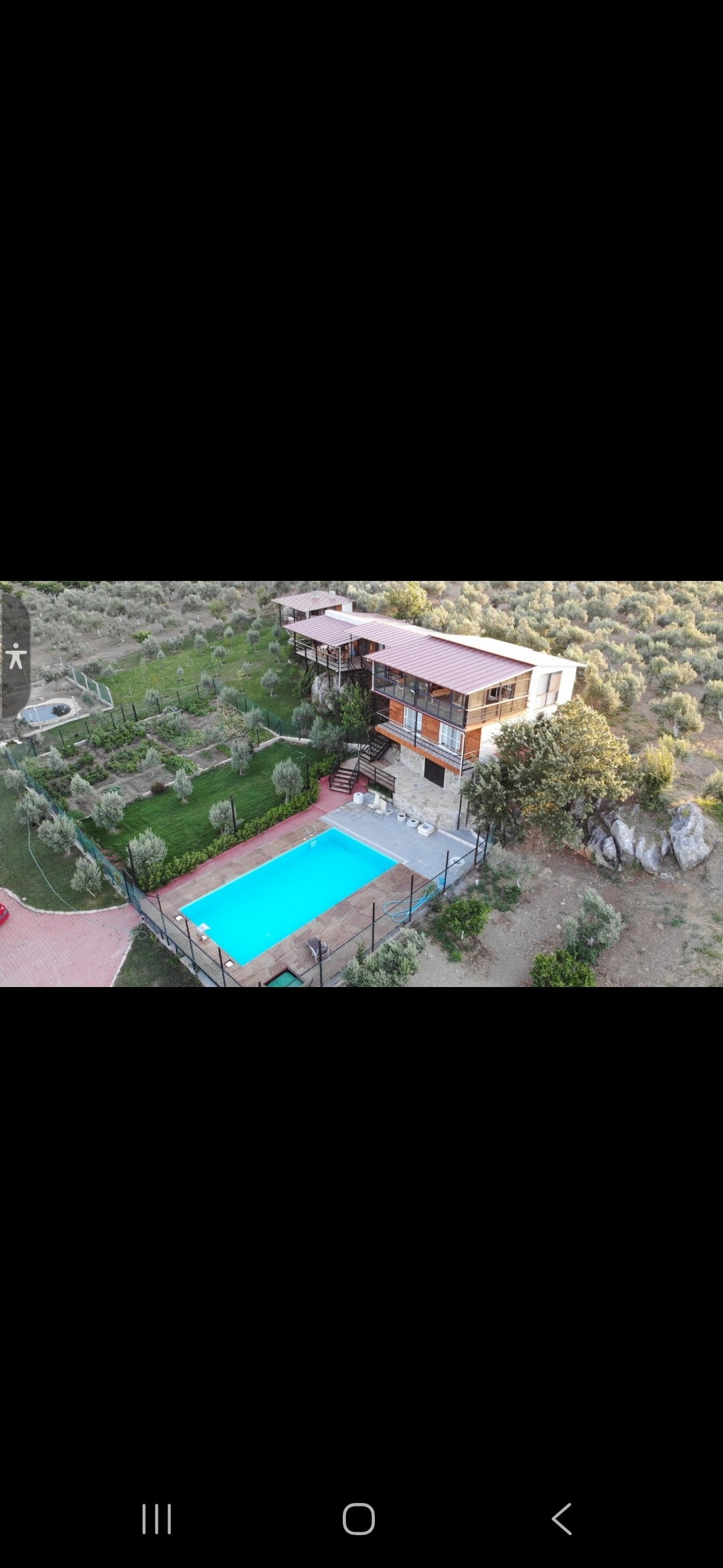 An aerial view captures a multi-level property surrounded by greenery. A turquoise pool is situated in the foreground, with sun loungers nearby. The structure features expansive balconies, framed by trees and landscaped gardens, indicating a serene outdoor environment.