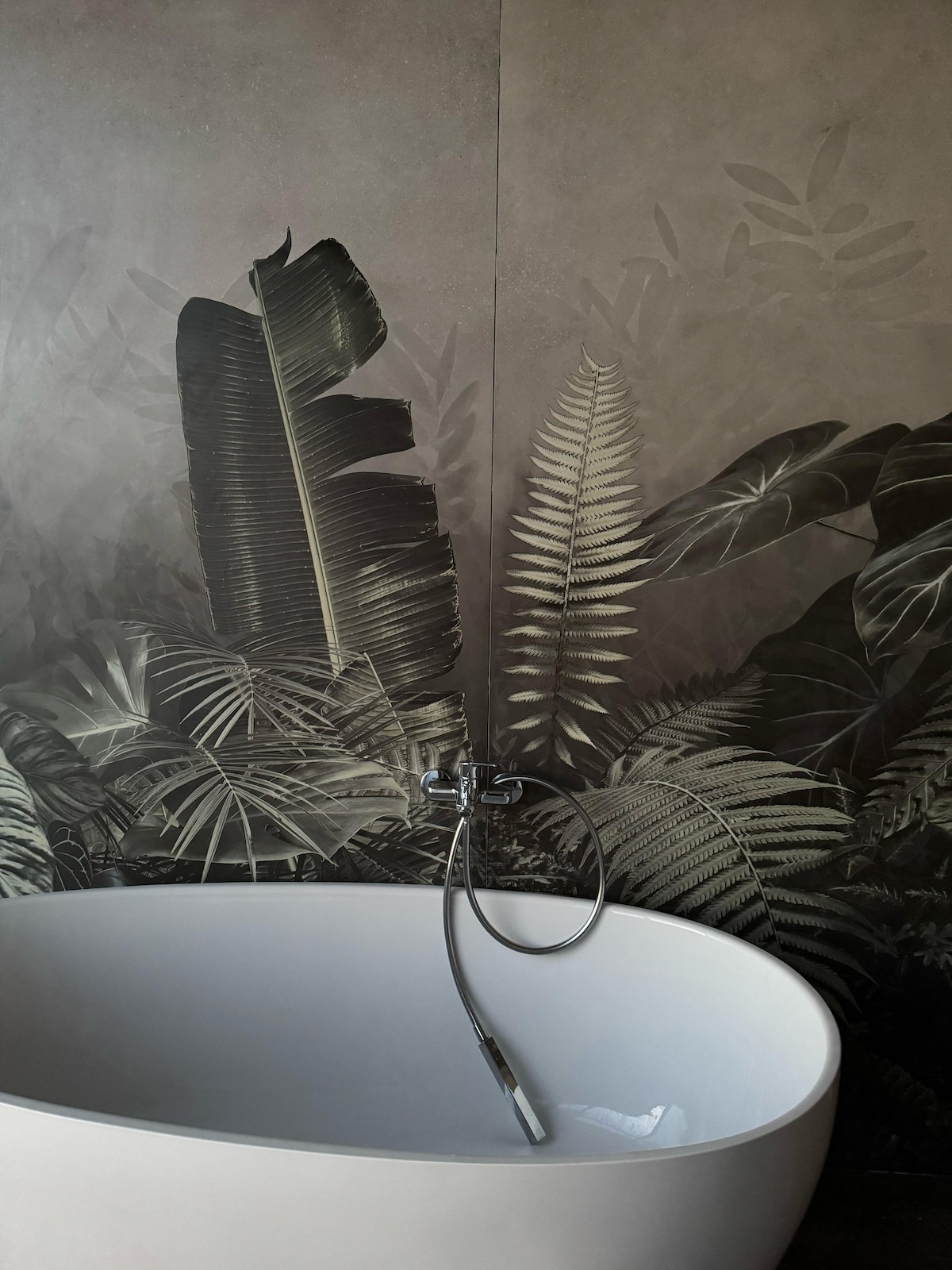 A modern freestanding bathtub is positioned against a mural of large, monochromatic ferns and leaves. The sleek, minimalist design of the bathtub is complemented by the understated elegance of the botanical artwork on the walls.