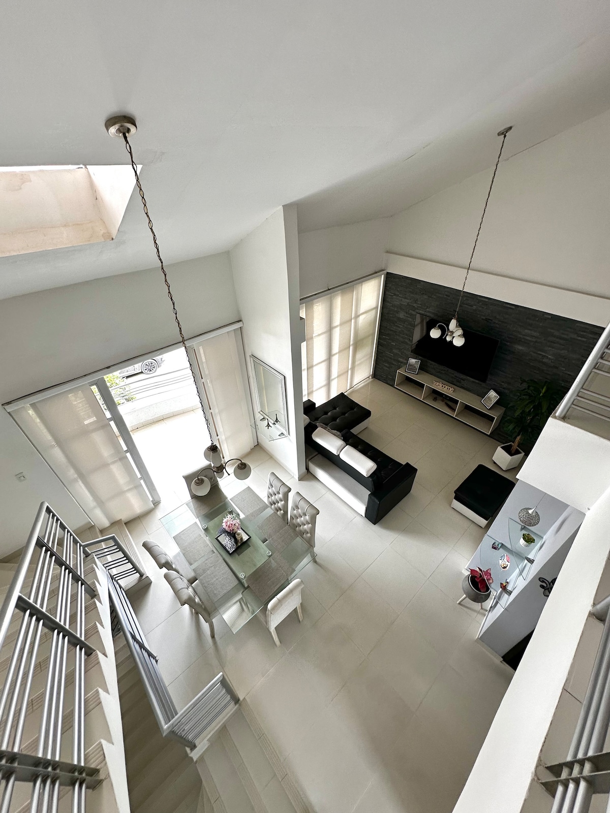 An aerial view captures the spacious living area bathed in natural light. A modern dining table is set for six, while comfortable seating is arranged nearby. Large windows allow sunlight to play across the room, emphasizing the minimalist decor.