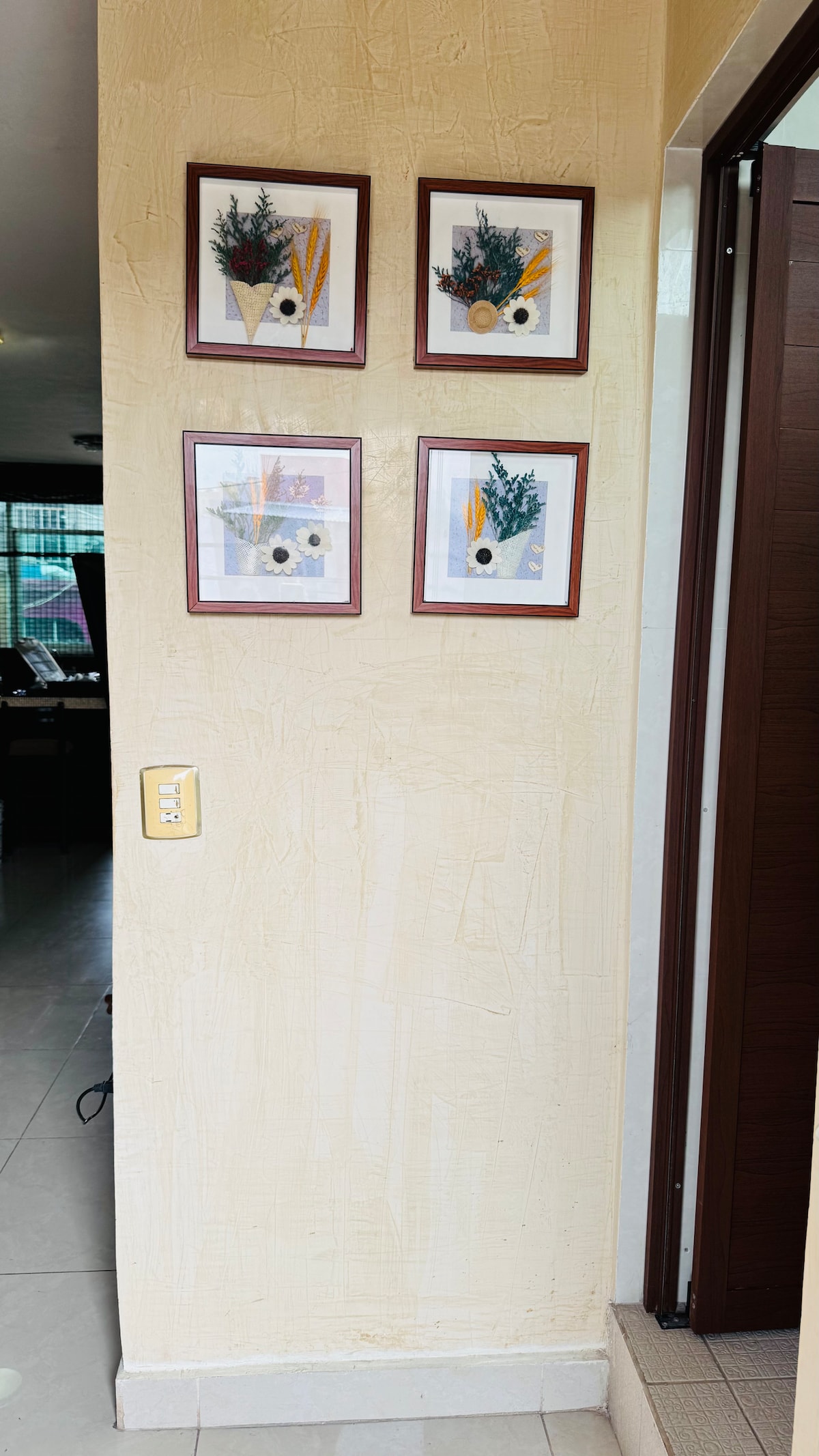 Four framed art pieces are arranged on a textured wall. Each piece features a combination of botanical illustrations and abstract elements. The warm tones of the frames complement the light color of the wall, creating a harmonious display near the entrance.