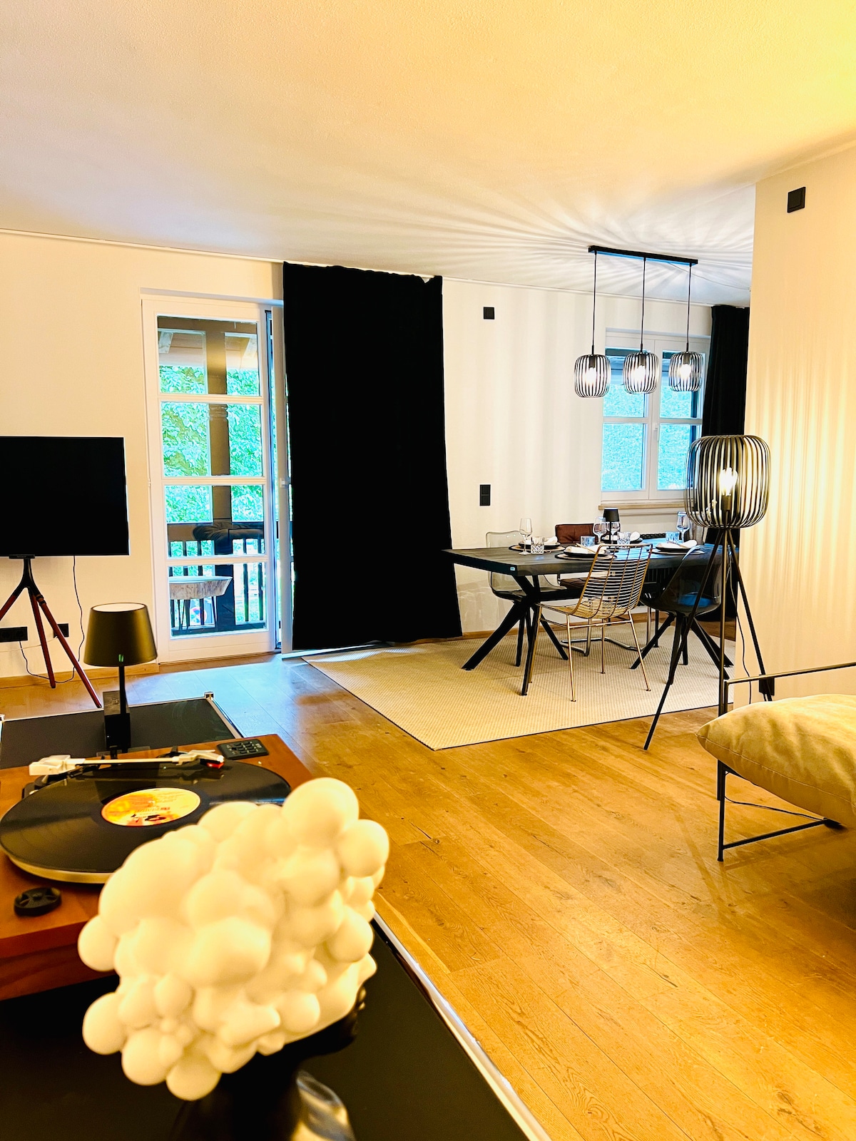 A living space is displayed with wooden flooring and ample natural light. A dining area is visible, featuring a dark table with transparent chairs. A black curtain contrasts with bright walls, while a record player and decorative elements add character to the room.