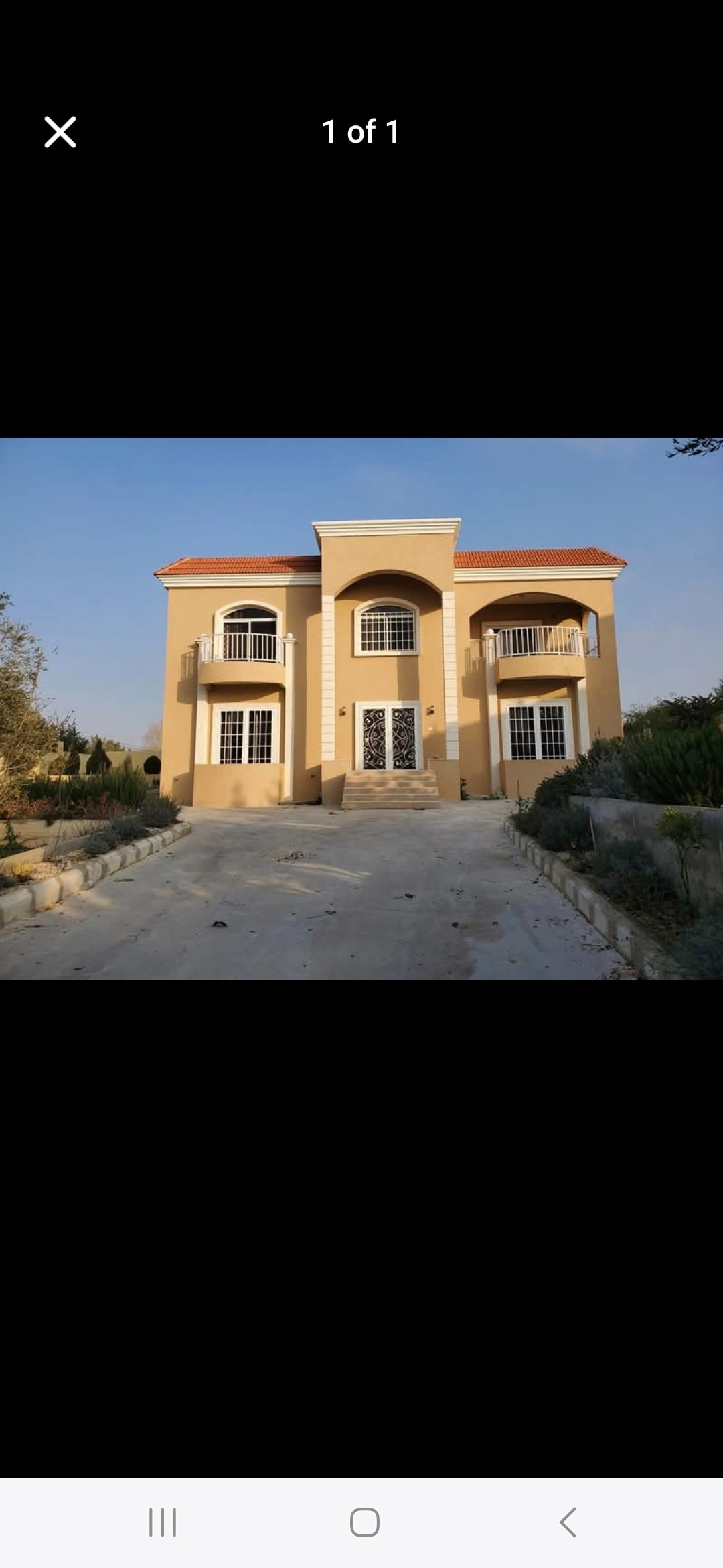 A grand two-story house displays a beige exterior with a red-tiled roof. Large arched entrances are framed by classic columns, while multiple windows invite natural light. The surrounding landscaped area features stone pathways and greenery, contributing to the property’s welcoming appearance.