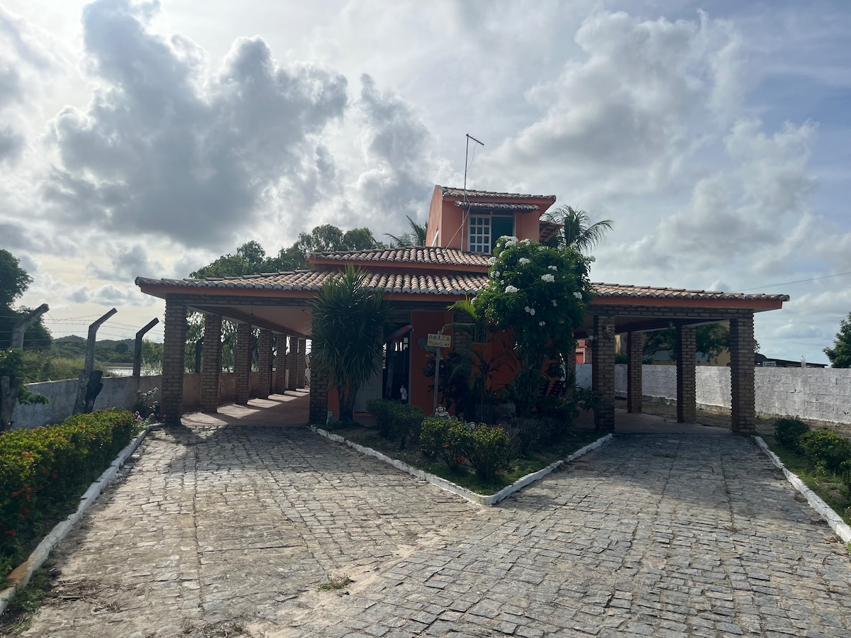 An inviting house with a terracotta roof is framed by lush greenery and a well-maintained garden. The entrance is approached via a cobblestone path, leading to a spacious veranda that extends around the front of the home, creating an open and airy environment.