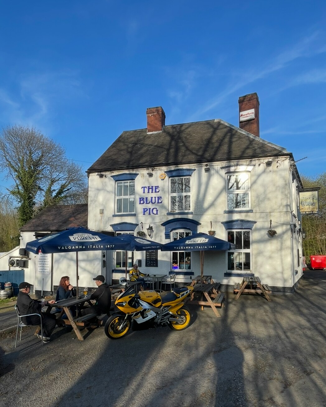 The exterior of The Blue Pig pub is shown, featuring a white façade and dark blue accents. Tables with umbrellas are seen outside, where guests are seated. A motorcycle is parked nearby, and trees cast shadows on the ground under the clear blue sky.