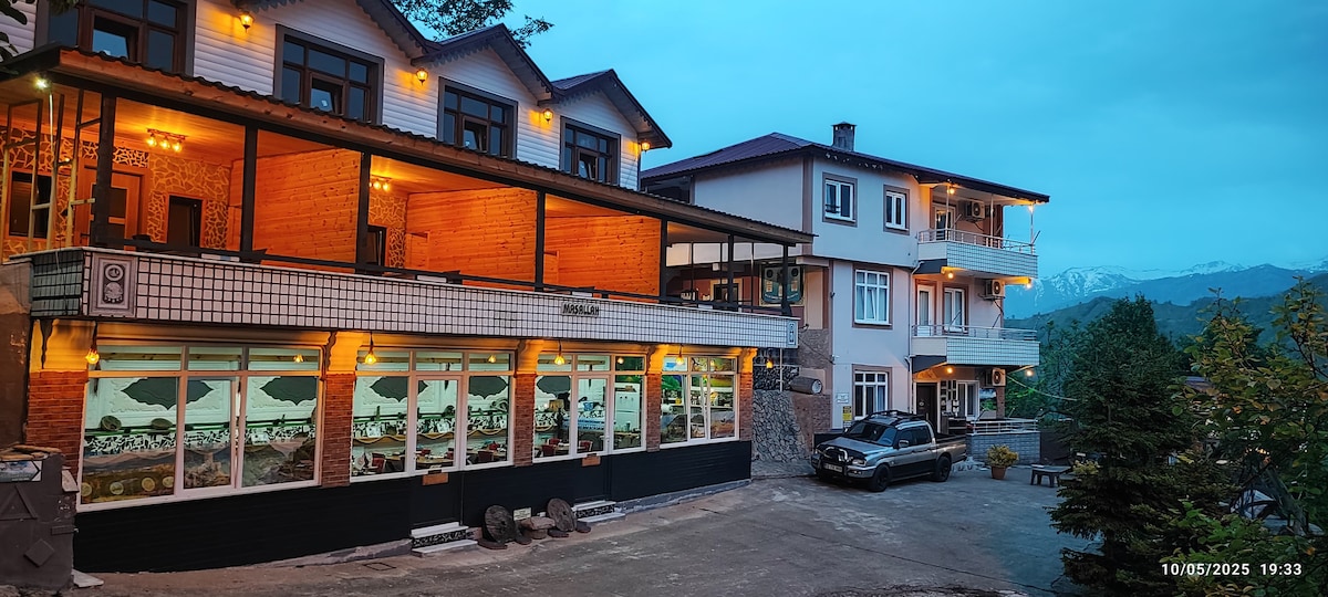 A two-story building with a combination of rustic wood and modern architecture is displayed in the early evening light. Soft illumination is seen along the exterior, accentuating the welcoming atmosphere. Ample outdoor space is visible, featuring a parked car and greenery in the background.