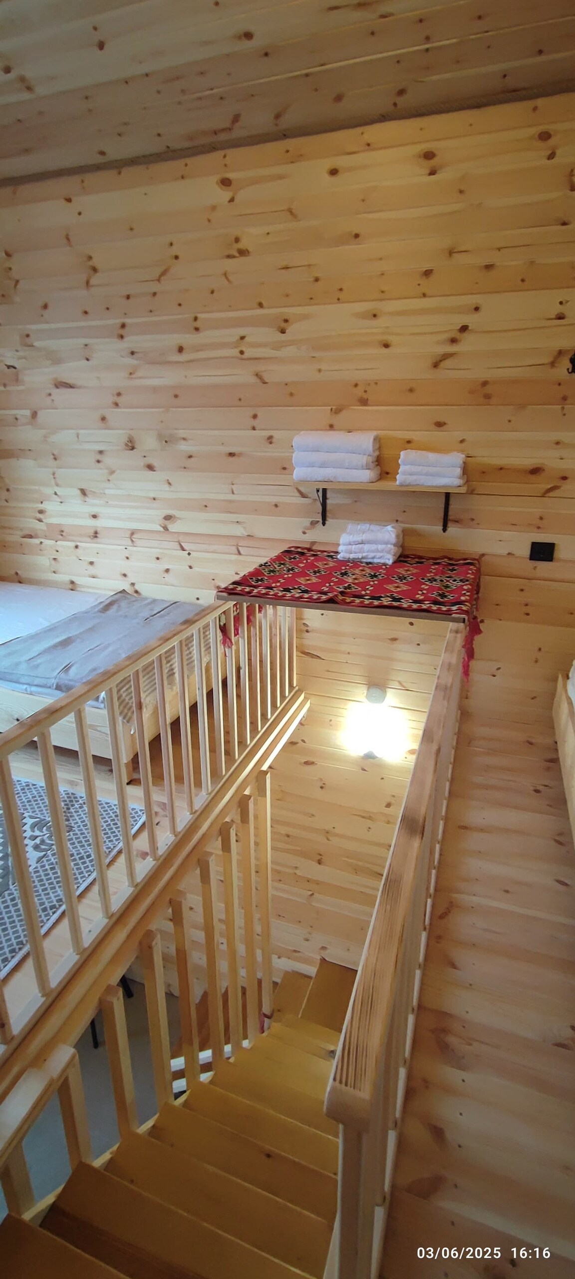 A wooden staircase leads to a loft area, showcasing natural wood paneling on the walls. Fresh white towels are neatly arranged on a shelf above a decorative textile, while a cozy sleeping area with beds is visible at the far end. Soft lighting enhances the warm ambiance.
