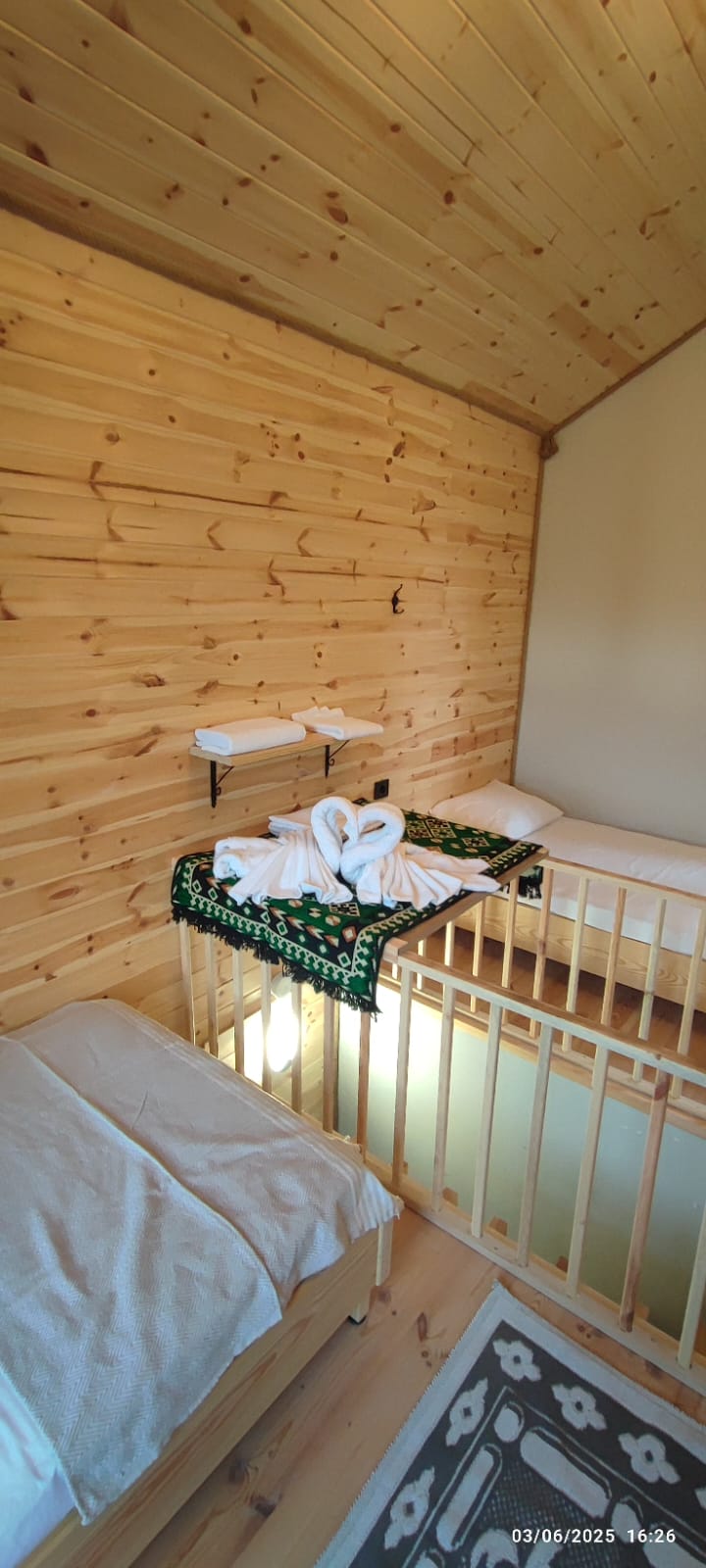 A room features wooden walls and flooring, providing a natural ambiance. Two beds are presented, one resembling a crib, adorned with neatly arranged towels shaped like swans. A decorative blanket lies across the crib, enhancing the space's inviting layout.