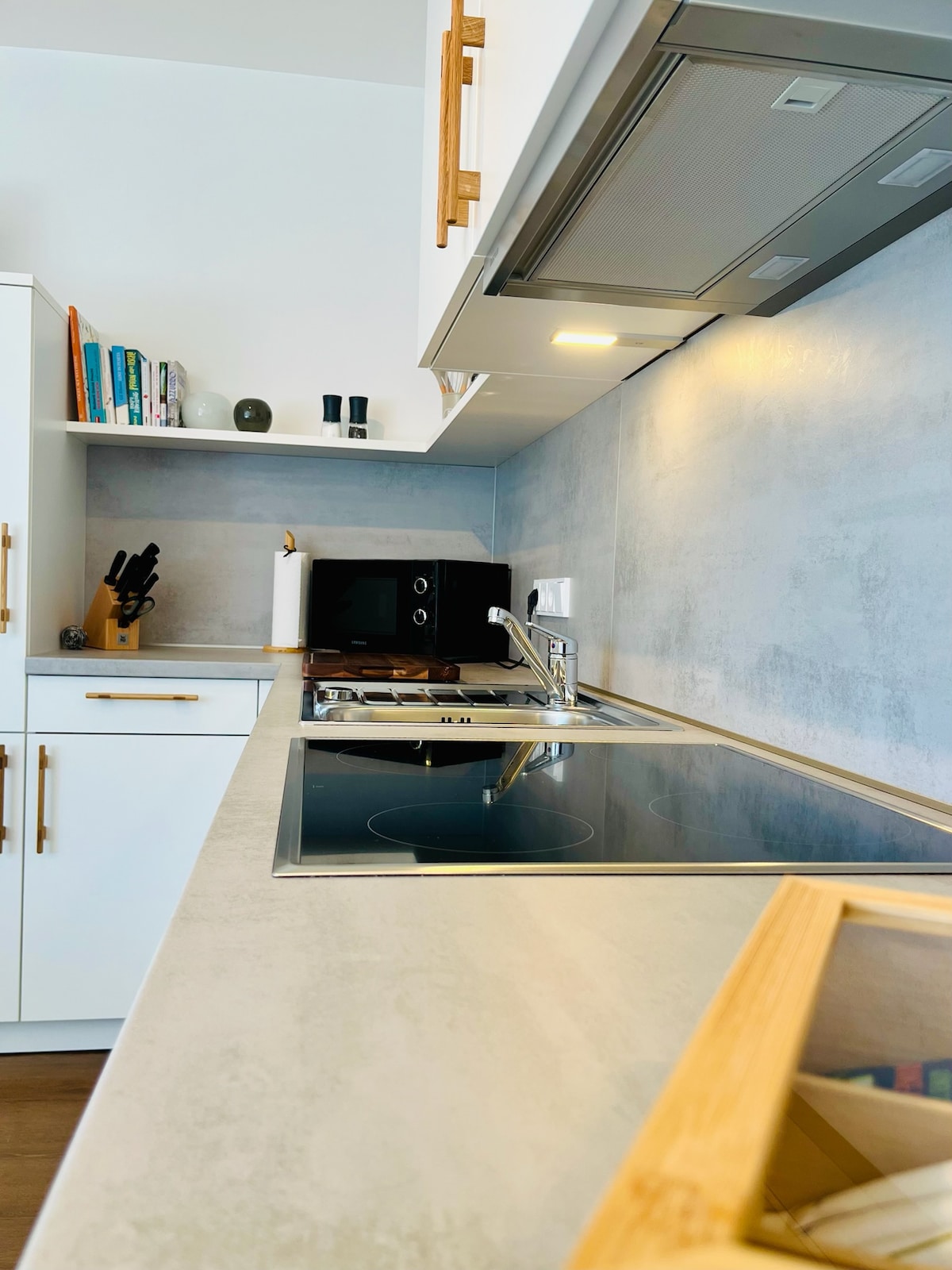 A modern kitchen workspace features sleek cabinetry and a light-colored countertop. An induction stove and sink are positioned along one side, with a microwave and minimalist decor items nearby. Utensils are neatly arranged, while shelves display books and decorative pieces.