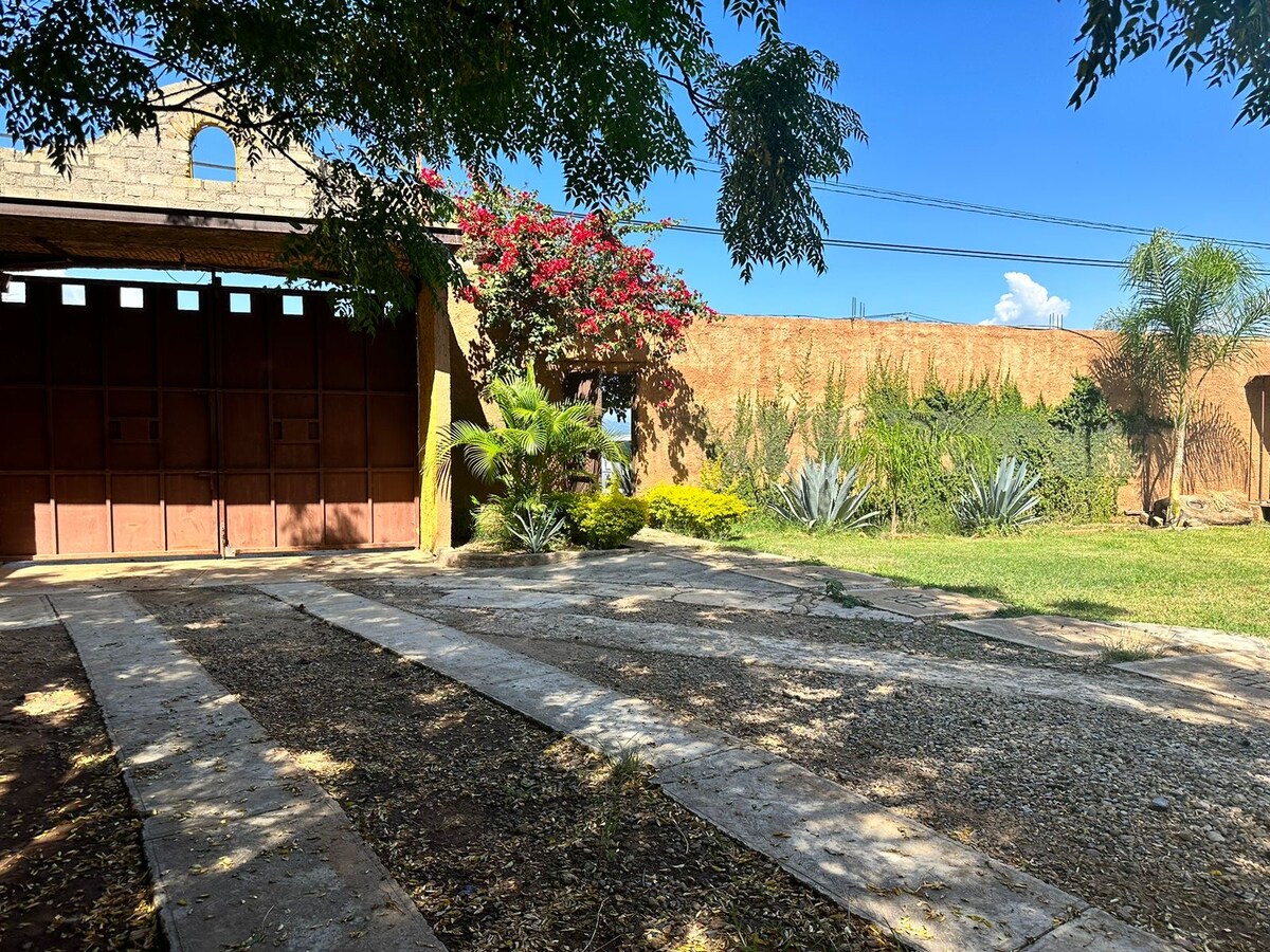 A gravel pathway leads to a wooden entrance gate, flanked by vibrant greenery and blooming flowers. The setting features a spacious outdoor area with a well-maintained lawn and various plants, creating a serene environment under a clear blue sky.