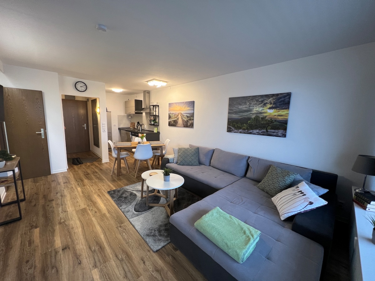 An open living area features a comfortable sectional sofa adjacent to a coffee table and a dining table set for four. Two landscape paintings adorn the walls, while a well-equipped kitchenette is visible in the background. Natural light fills the space through a window.