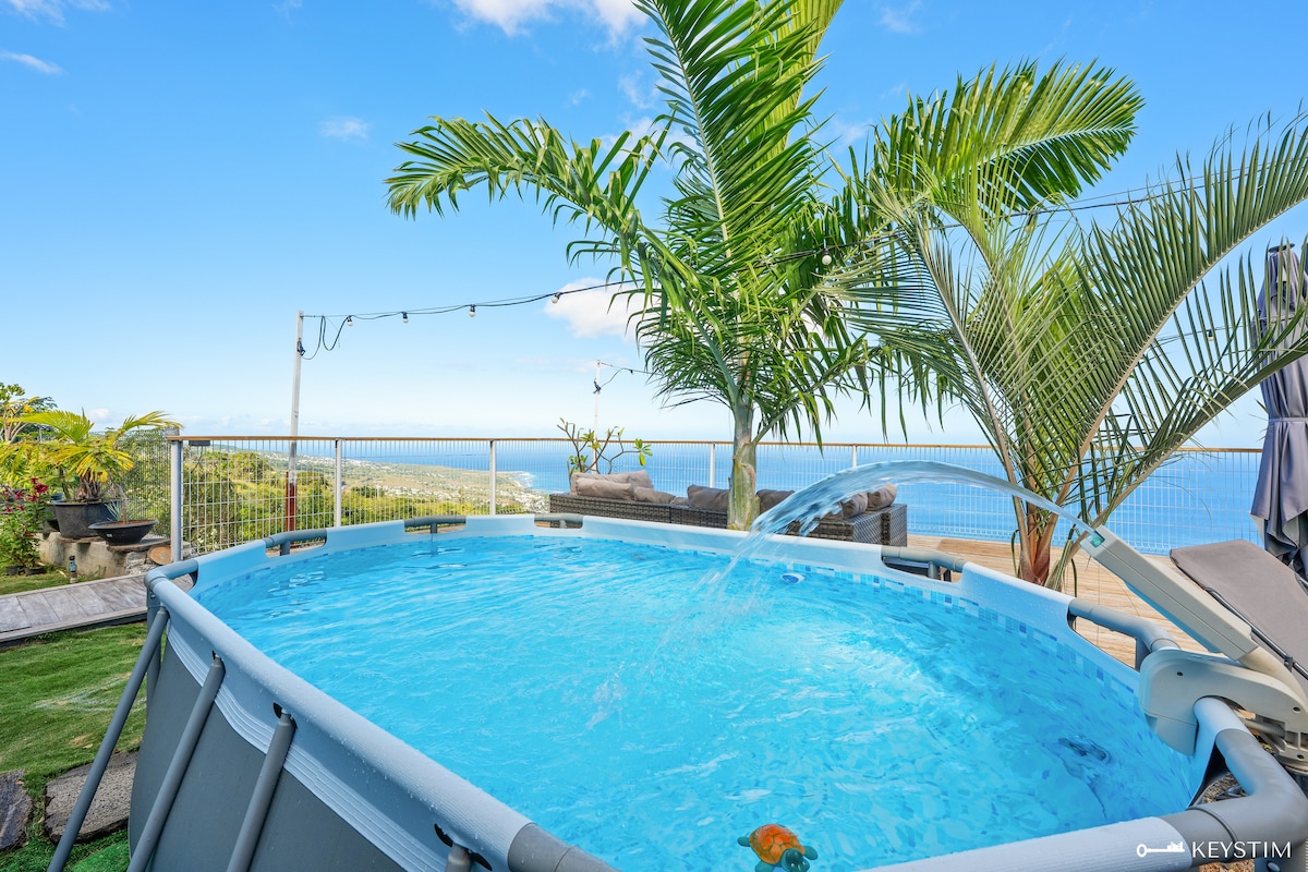 A private above-ground pool is surrounded by lush greenery, including palm trees. The clear water reflects the blue sky, and a gentle fountain feature adds movement. A panoramic view of the ocean is visible in the background, enhancing the serene outdoor atmosphere.