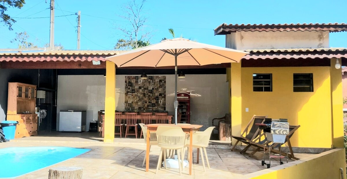 An outdoor area features a swimming pool surrounded by a spacious patio. Dining furniture is set under a large umbrella. The structure showcases a mix of yellow walls and tiled accents, with an integrated barbecue space and a kitchen visible in the background.