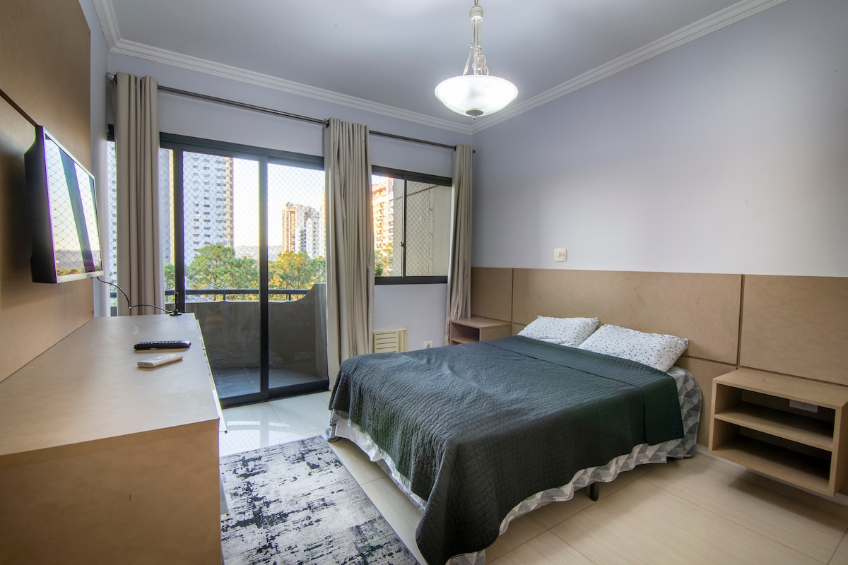 A comfortable bedroom is furnished with a spacious bed covered in a dark quilt. The room features light-colored walls and a large window that offers views of the city outside. A TV is mounted on the wall opposite the bed, and light drapes frame the window.