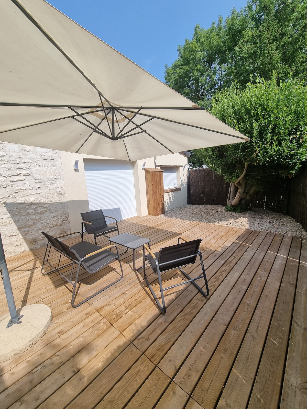 A spacious terrace is depicted, featuring a wooden deck and three lightweight chairs arranged beneath a large umbrella. Surrounding greenery provides a sense of privacy, while a smooth stone path leads to a gated area. Natural light fills the space, enhancing the inviting outdoor setting.