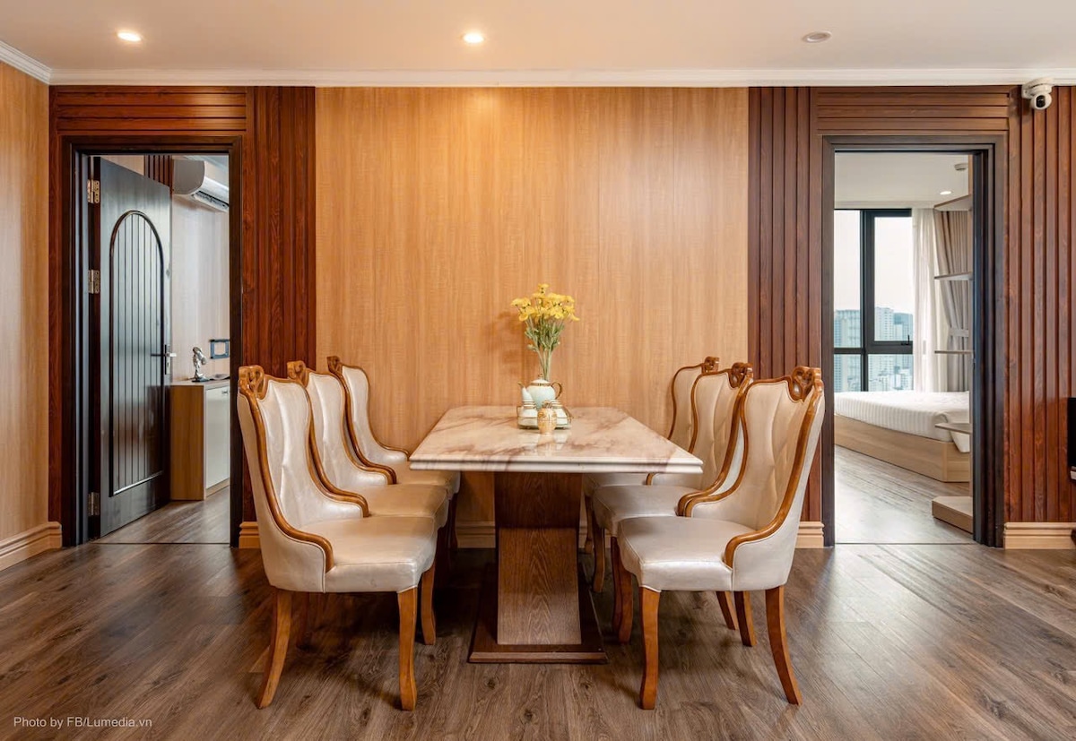 A dining area features a rectangular marble table surrounded by six upholstered chairs with elegant wooden accents. Light tones of the wood paneling create a warm backdrop, while a small vase of flowers sits at the center of the table, adding a touch of charm to the space.