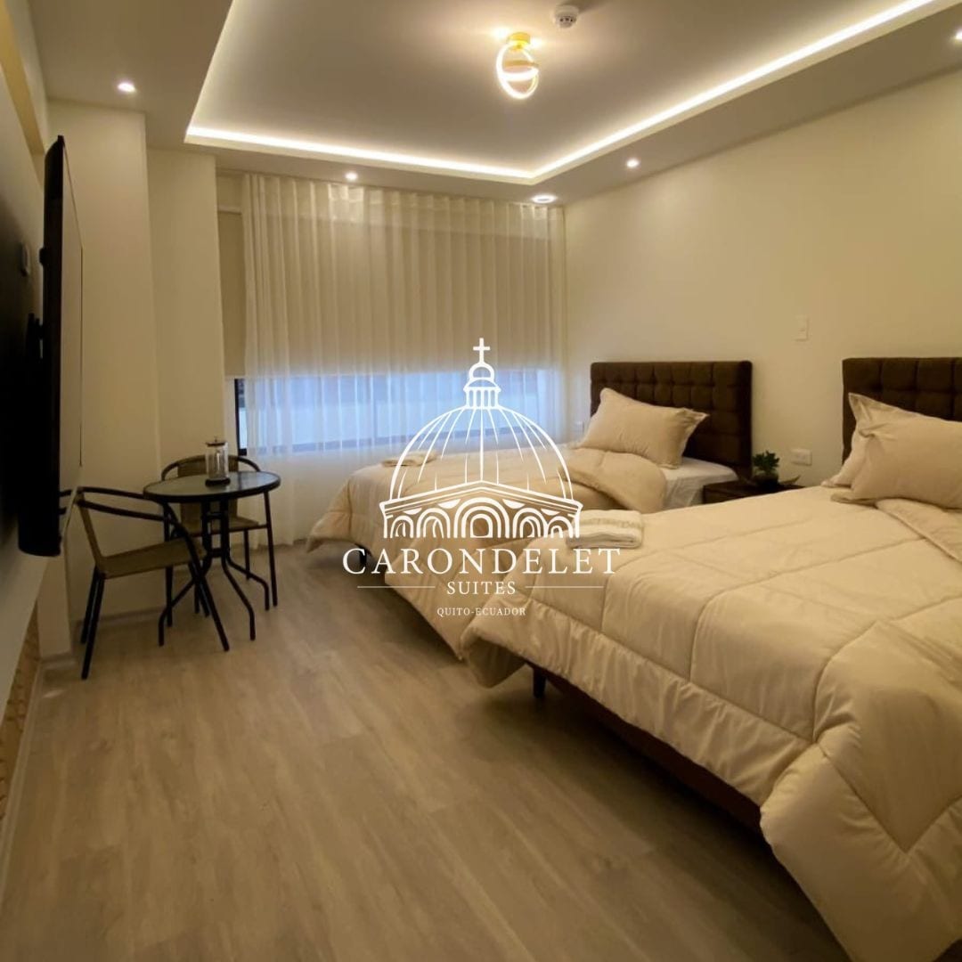 The room features two spacious beds with neutral-colored bedding, complemented by a modern wall-mounted television. A small table and chair set is positioned by the window, allowing for natural light to enter the space. Soft, ambient lighting is provided by recessed ceiling fixtures.