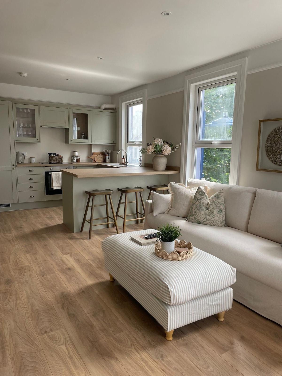 An open-plan living and dining area features light wooden flooring and large windows allowing natural light. A comfortable sofa is paired with a striped ottoman, and a well-equipped kitchen is visible in the background, showcasing modern appliances and a bar area with two stools.
