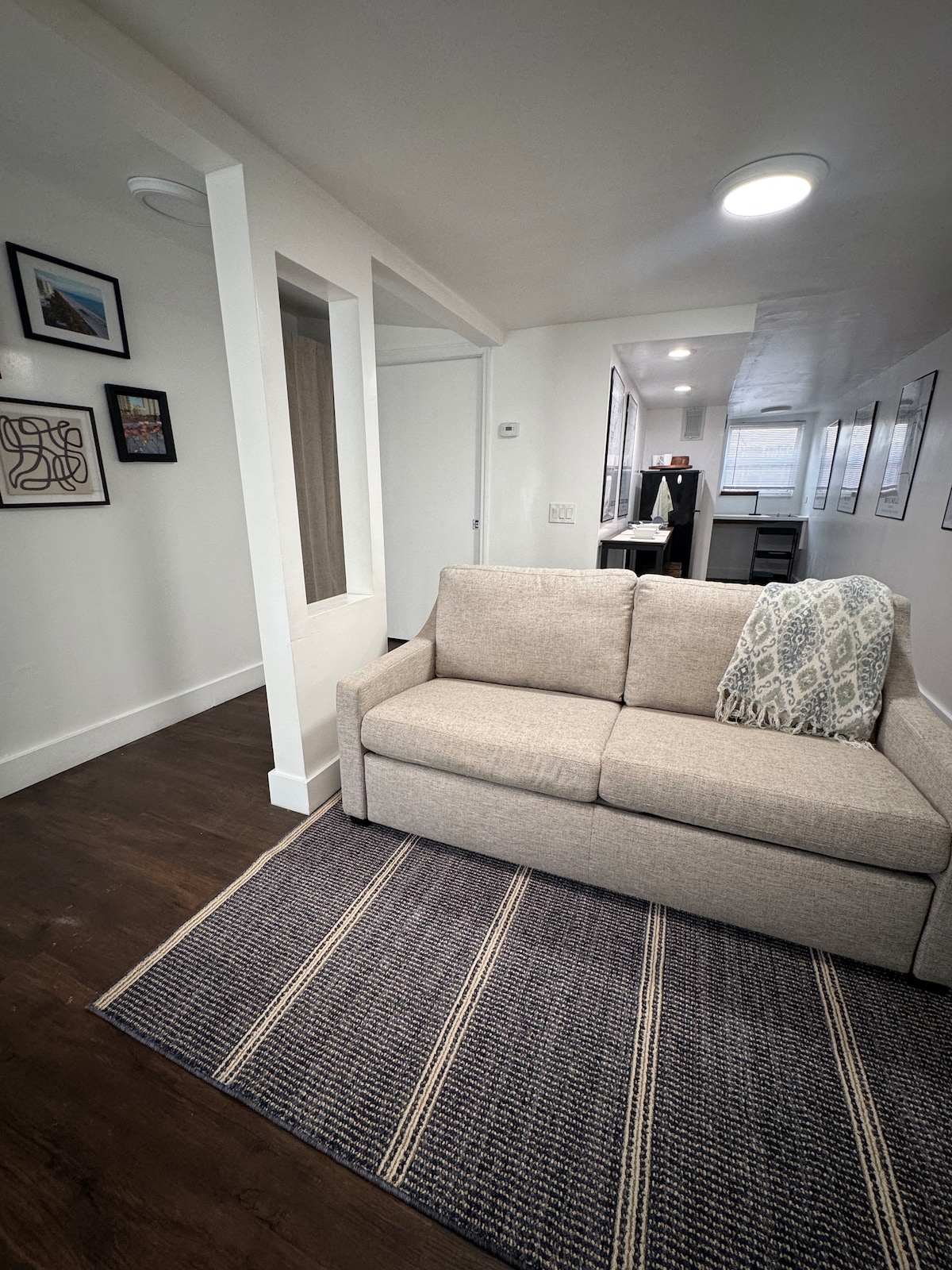 A light-filled living area showcases a cozy sofa with a decorative throw pillow. A patterned area rug anchors the space, while framed artwork adorns the walls, adding a touch of character. A doorway is visible, leading to other functional areas of the retreat.