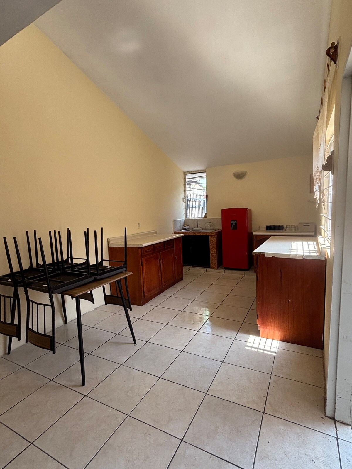 A light-filled kitchen features light-colored walls and tiled flooring. Wooden cabinets and a dark countertop provide storage and workspace. A red refrigerator stands prominently against one wall, while a set of chairs is stacked on a table, indicating a spacious and functional environment.