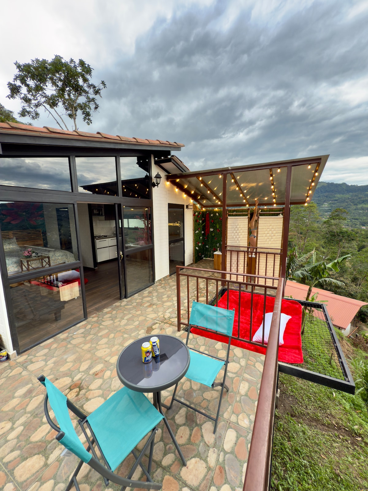 An outdoor terrace is shown, featuring a small table with two turquoise chairs. A red blanket is laid out on the grass, and string lights hang above, illuminating the area. The backdrop includes lush greenery and distant hills under an overcast sky.