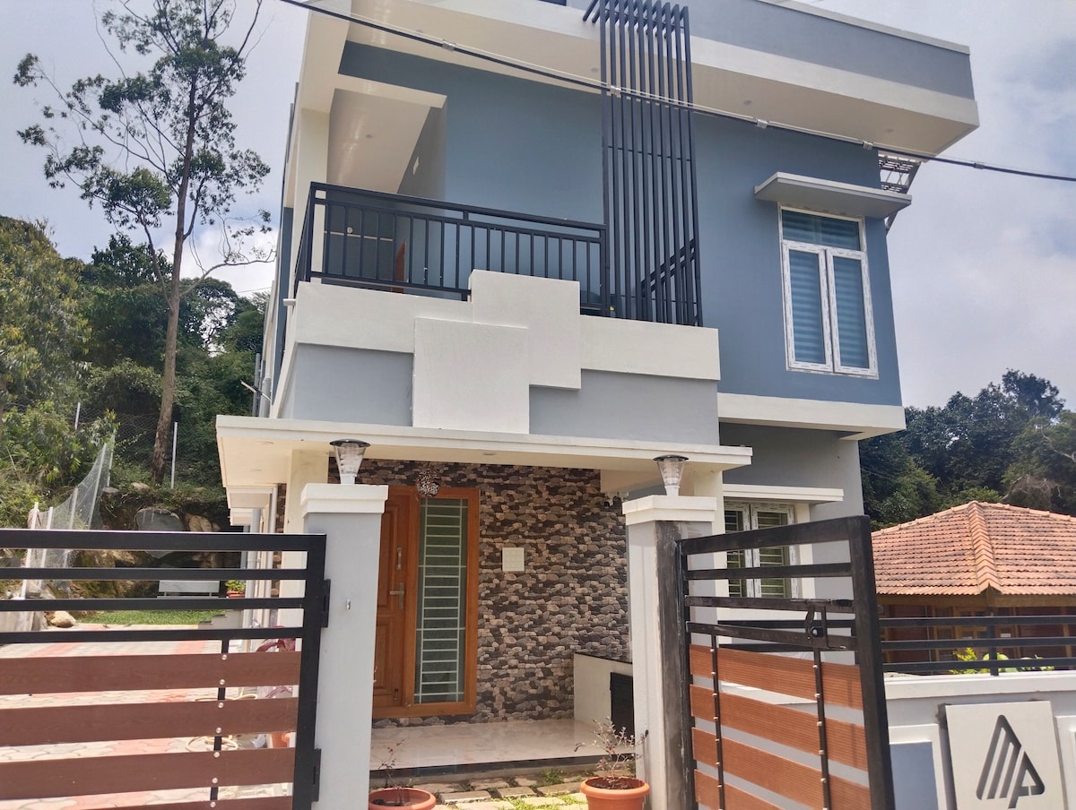 The exterior of a modern villa is showcased, featuring a combination of stone and painted surfaces. A balcony extends from the upper floor, bordered by sleek railings. A gated entrance leads into a small garden area, with trees and neighboring buildings visible in the background.