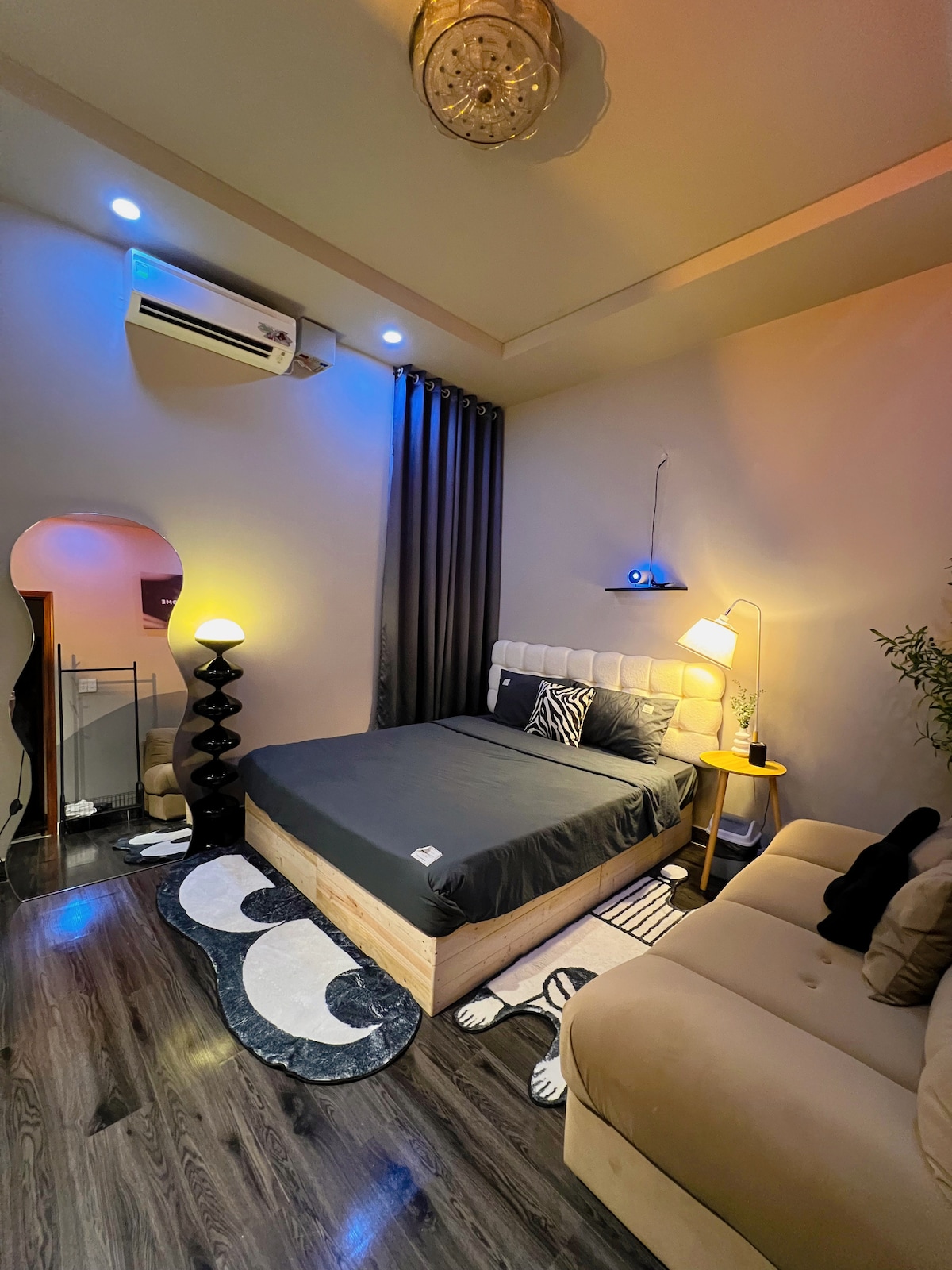 A cozy room features a spacious bed with dark bedding, complemented by a decorative headboard. Soft lighting creates a warm atmosphere, while a comfortable sofa and stylish black floor lamp add to the soothing decor. Curtains frame a window, enhancing the room's inviting feel.