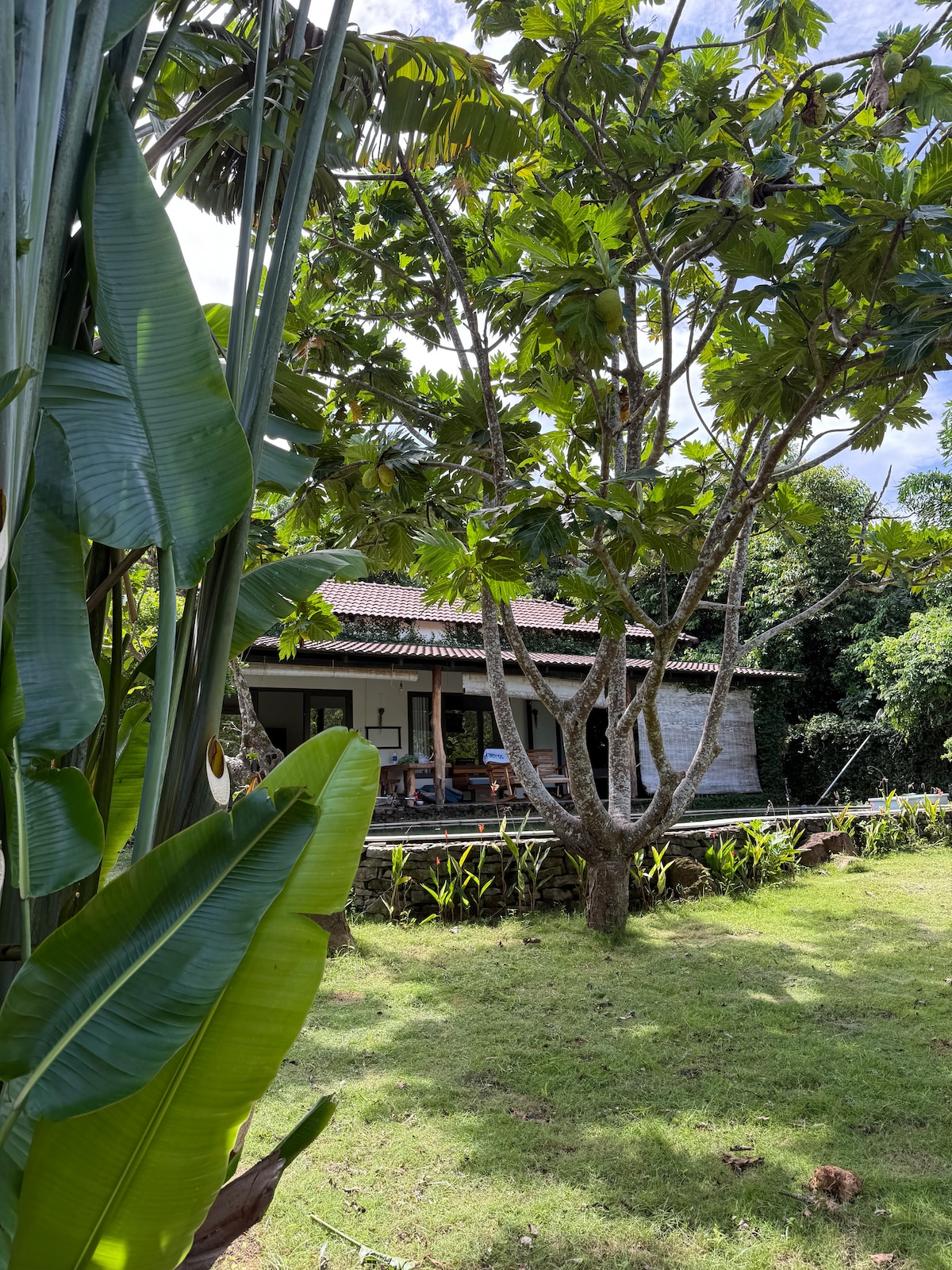 A charming villa is set among lush greenery, featuring a rustic exterior and a welcoming porch. Large trees provide shade, and the surrounding grassy area invites relaxation. Visible elements include a comfortable seating arrangement on the porch, blending harmoniously with the natural surroundings.