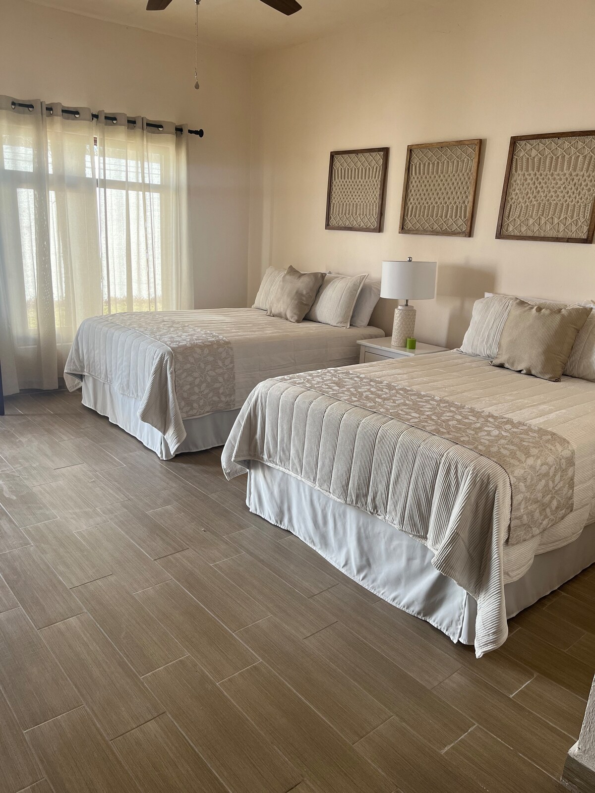 The sleeping area is furnished with two spacious beds, each topped with light-colored bedding. Nightstands with lamps are positioned beside the beds. Natural light filters through sheer curtains, illuminating the room's neutral tones and simple wall art.