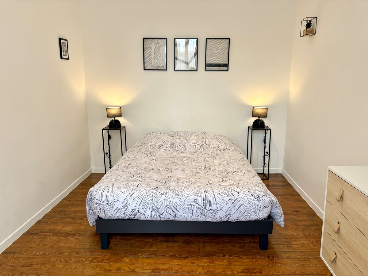 The bedroom features a spacious bed dressed in a patterned duvet cover. Wall-mounted lamps provide soft illumination on either side, complemented by framed artwork above the bed. A light-colored dresser is positioned nearby against the wall, enhancing the room's functionality.