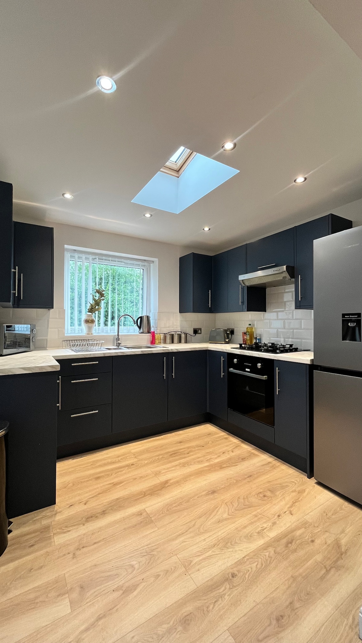 A modern kitchen is equipped with sleek navy blue cabinetry and stainless steel appliances. Natural light enters through a skylight above the sink, illuminating the workspace. A neutral countertop contrasts with the dark cabinets, while light-colored flooring complements the overall design.