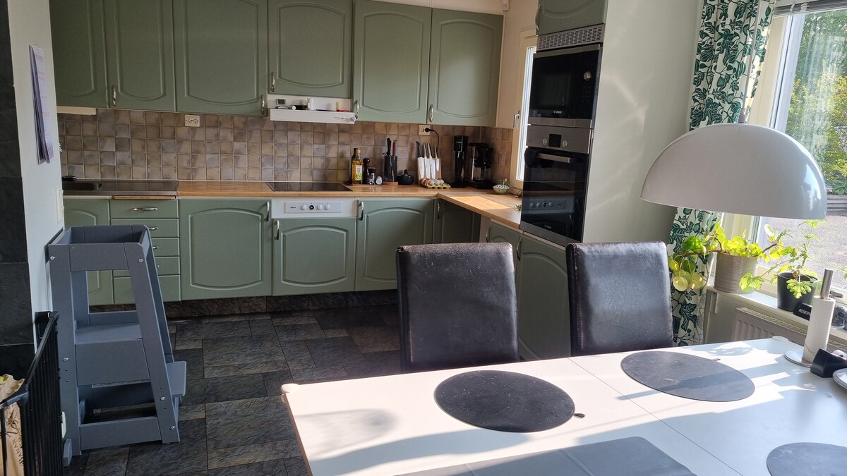The kitchen showcases an array of green cabinets and a tiled backsplash. A wooden countertop runs between the cabinets and built-in appliances, including an oven. A dining table is set with placemats, surrounded by several black chairs, while natural light filters through a large window.