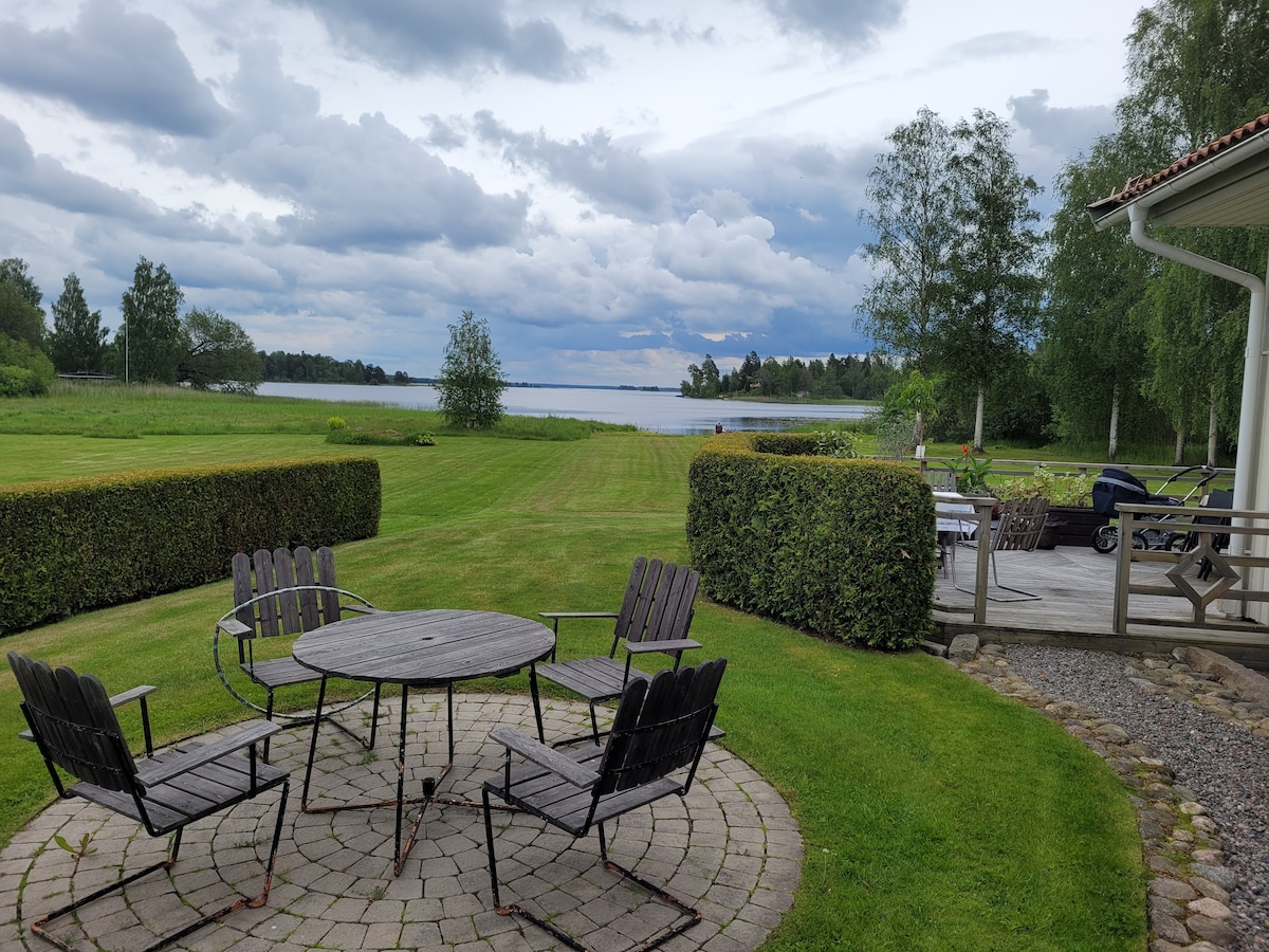 A circular stone patio is surrounded by six wooden chairs and a central table. Lush green grass extends towards a serene lake, with trees lining the shore under a cloudy sky. A spacious deck is visible to the right, complementing the outdoor setting.