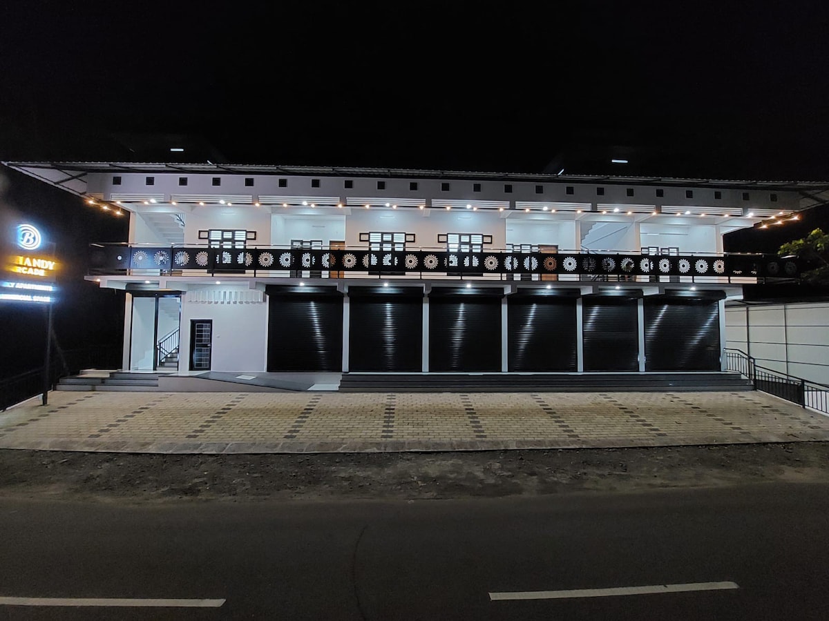 The building features a modern design with a clean façade and multiple windows. It is well-lit, highlighting an expansive entrance area and decorative elements along the balcony. The surrounding area has been paved with a light-colored stone, providing a clear path for entry.