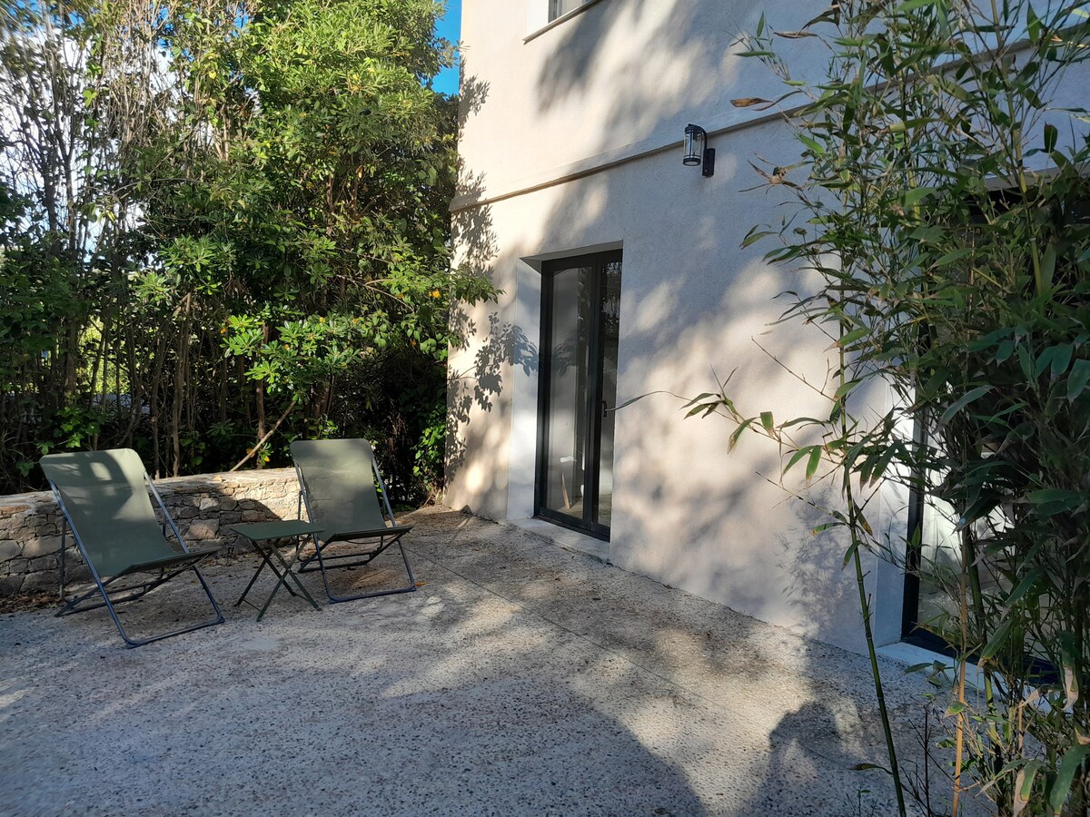 A small outdoor terrace features two green reclining chairs positioned on a gravel surface. Sunlight filters through nearby foliage, creating a mix of light and shadow on the wall of the building. A large window is visible, enhancing the connection between indoor and outdoor spaces.