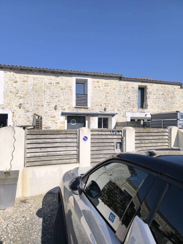 A charming stone building is depicted with a clear blue sky above. The facade features white windows and a wooden railing, with an adjacent gray car parked in front. A fenced area is visible, highlighting the inviting exterior of the property.