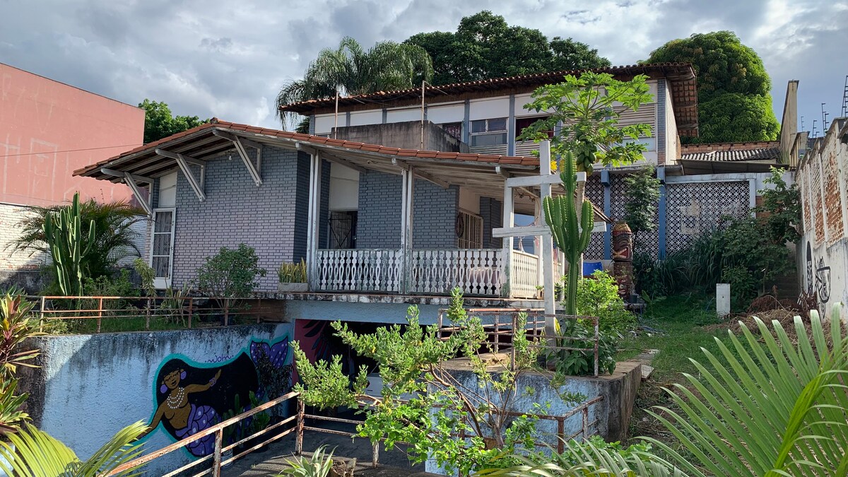 A multi-level building is surrounded by lush greenery, featuring a mix of modern and traditional architectural elements. A wide deck is seen in the foreground, alongside colorful murals and a gently sloped garden area, providing a vibrant outdoor atmosphere.