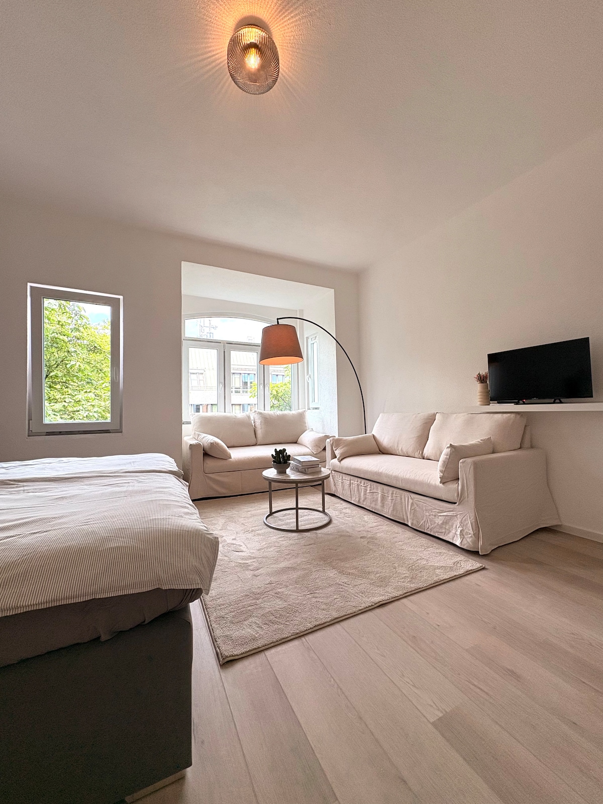 A light-filled living area features a soft beige sofa and a matching loveseat positioned around a small circular coffee table. Two windows offer views of greenery outside, while a modern floor lamp provides additional lighting. A TV is mounted on the wall opposite the seating.