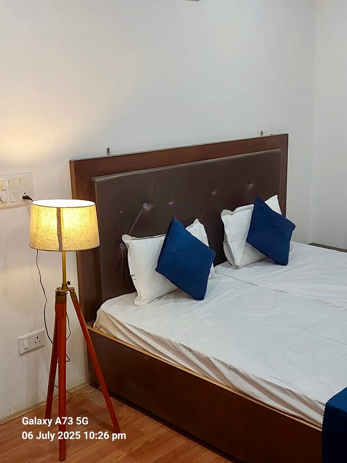 A cozy bedroom features a spacious bed adorned with white linens and dark blue accent pillows. A wooden floor complements a standing lamp with a beige shade, providing soft illumination. The minimalistic decor creates a serene sleeping environment.