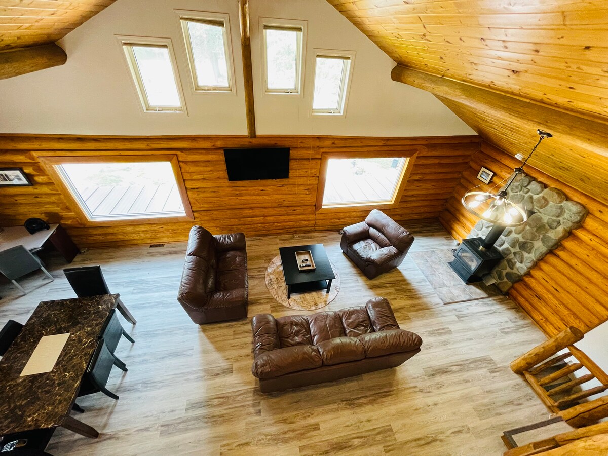 An open-concept living and dining area features vaulted wood ceilings and large windows that allow natural light to fill the space. Two brown leather sofas are positioned around a coffee table, with a dining table visible in the background. A stone fireplace adds rustic charm.