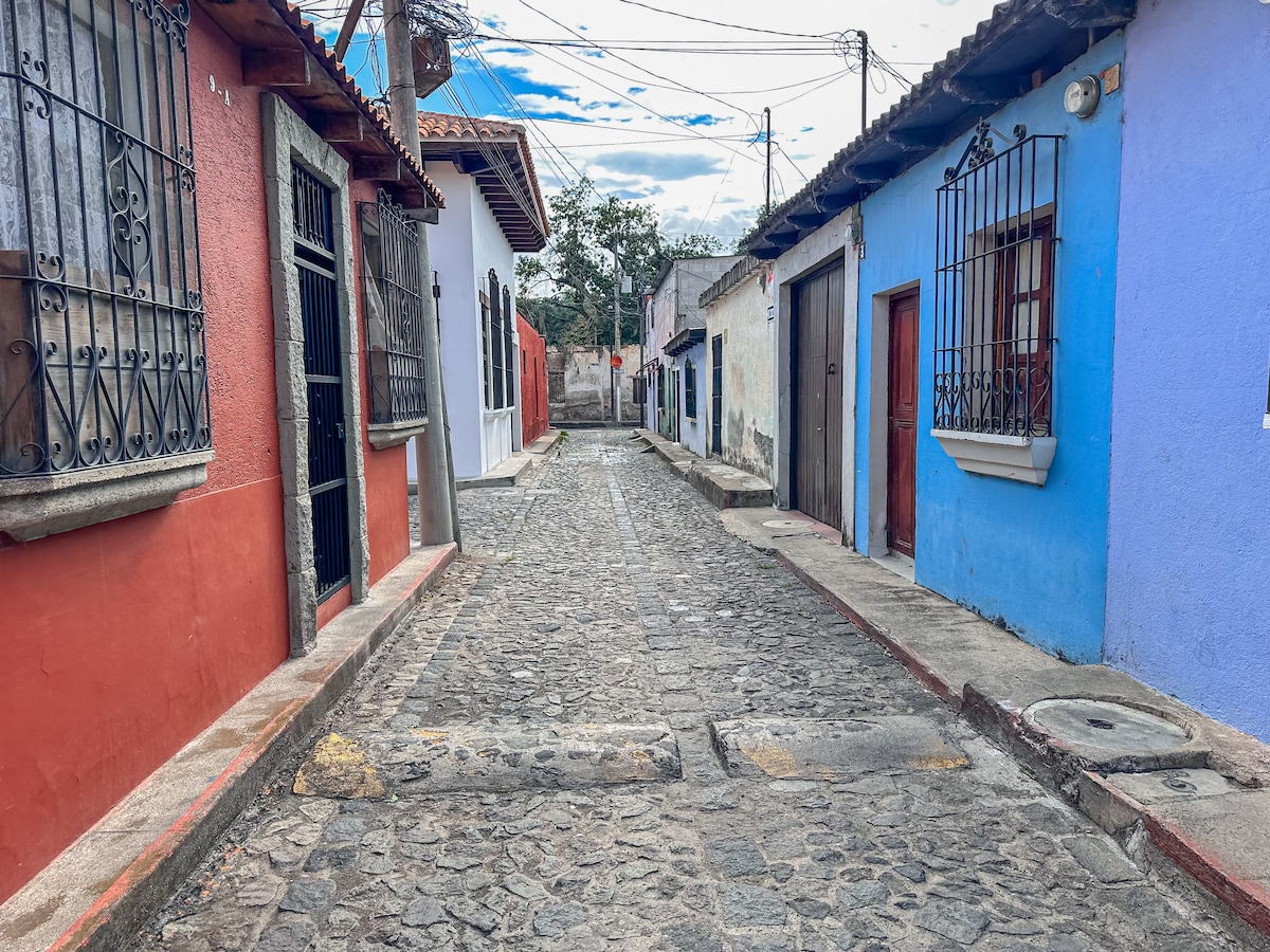 A charming cobblestone street is flanked by vibrant buildings in shades of red, white, and blue. Traditional windows with wrought-iron bars add character, while power lines stretch across the sky, hinting at the lively atmosphere of the surrounding neighborhood.