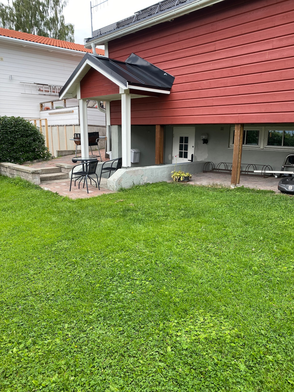 A well-maintained outdoor area features a patio with four black chairs and a small table under a roofed section. Lush green grass surrounds the space, while a pathway leads to a neighbouring building. An outdoor barbecue area is visible in the background.