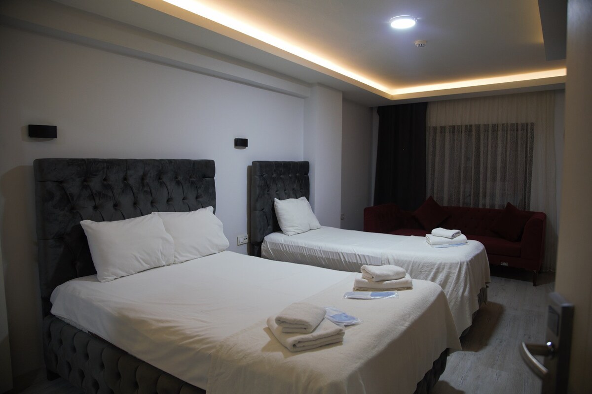 A well-appointed sleeping area is featured, showcasing two spacious beds with white linens and neatly arranged towels. A modern sofa adds additional seating, while soft lighting creates a calming ambiance. Large windows with light-colored curtains allow natural light to brighten the room.