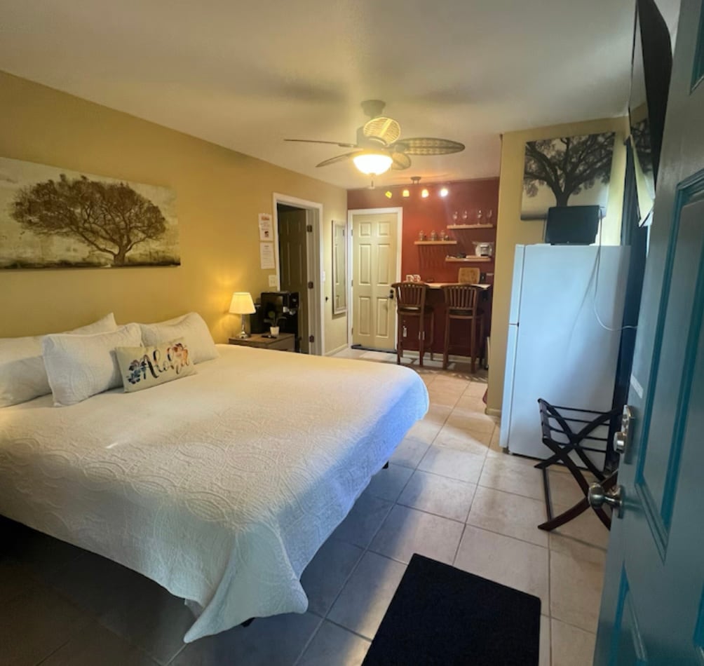 A spacious rental suite features a king bed with a white quilt and decorative pillows. A kitchenette is located in the background, complemented by a dining area with two bar stools. Natural light illuminates the room from windows, and a ceiling fan circulates air peacefully.