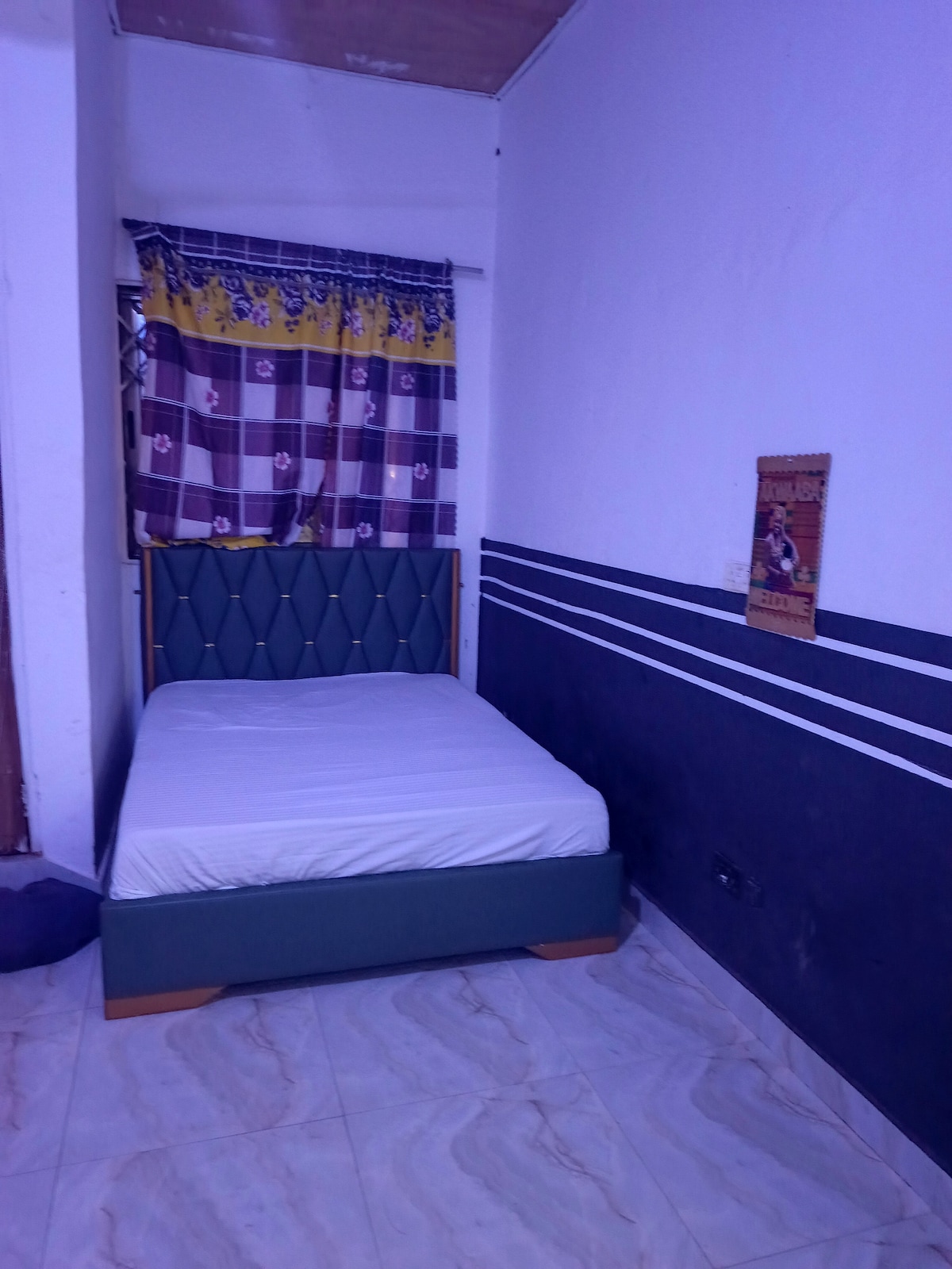 A simple sleeping area features a spacious bed with a green upholstered headboard against a pale wall. A colorful patterned curtain hangs over a nearby window, while the floor is covered with large, light-colored tiles.